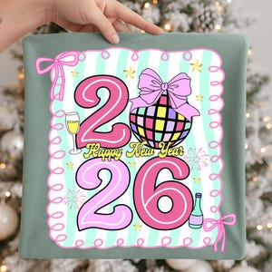 May include: A decorative pillow with a festive design for the New Year. The design features the numbers "26" in pink, a disco ball, champagne, and the words "Happy New Year". The pillow has a light green border and pink bows.
