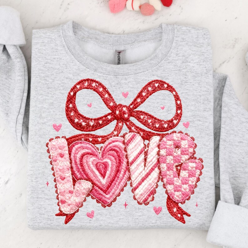May include: A light gray sweatshirt with a red bow and the word "LOVE" embroidered in pink and red. The "O" is a heart shape. Small pink hearts are scattered around the design. The sweatshirt is a crew neck style.