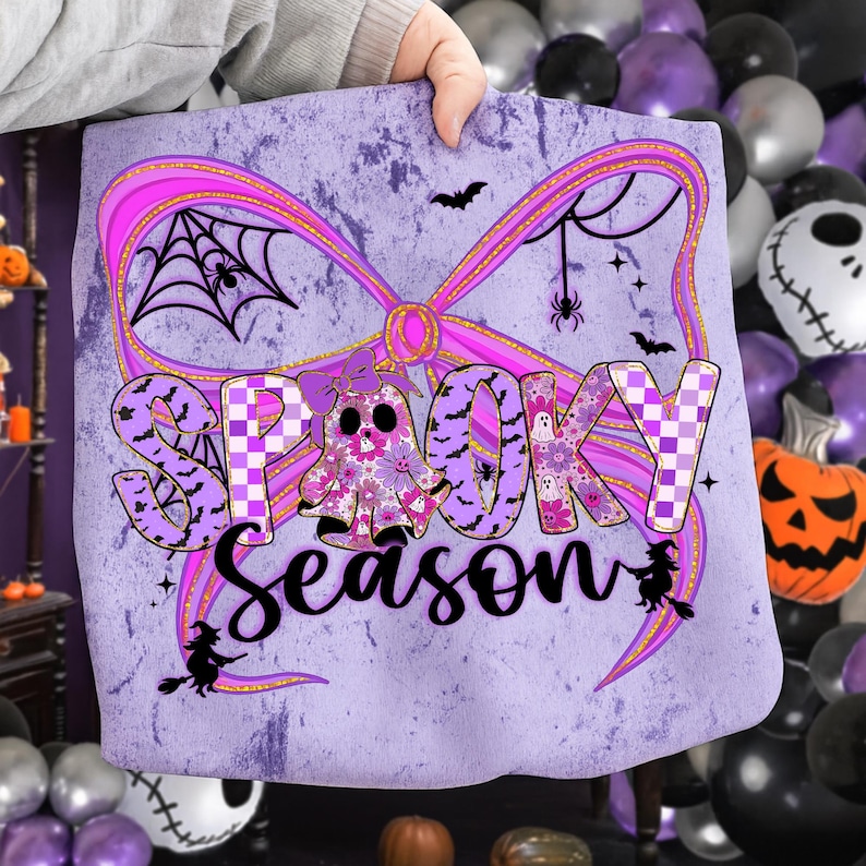 May include: A lavender tote bag with a Halloween theme. The design features a large pink bow, the words "Spooky Season", bats, spiders, and a ghost with floral patterns. The background has a distressed texture.