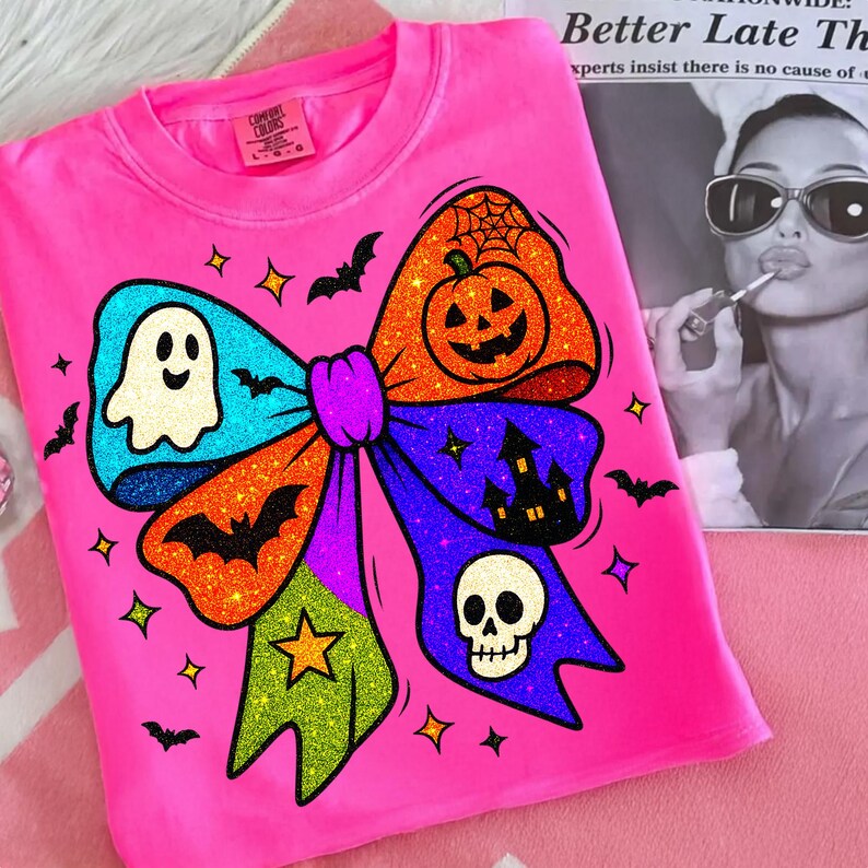 May include: A bright pink sweatshirt featuring a large, glittery Halloween bow design. The bow includes a ghost, bats, a pumpkin, a haunted house, a skull, and stars. The sweatshirt is a Comfort Colors brand.