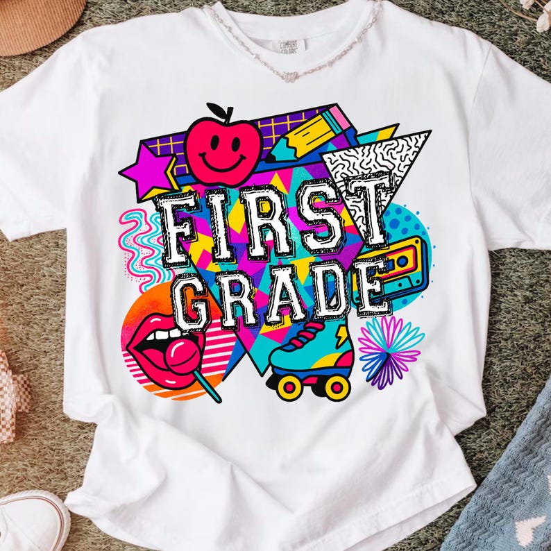 Pu&ograve; includere: T-shirt bianco con grafica colorata "FIRST GRADE". Il design include una mela sorridente, un pattino a rotelle, una cassetta e un lecca-lecca. Il design ha uno stile retr&ograve; anni '80 e '90.