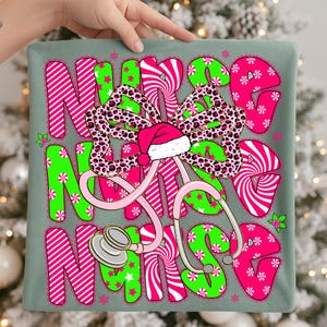 May include: A decorative pillow with the word "NURSE" in pink and green, featuring a stethoscope and a Santa hat. The letters have a candy cane and star pattern. A leopard print bow is in the center. The pillow is held by a hand.