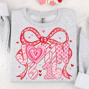 May include: A light gray sweatshirt with a large red bow and the text "JOHN 3:16" in pink and white. The design includes hearts and a heart-shaped candy. The sweatshirt is a Valentine's Day themed item.
