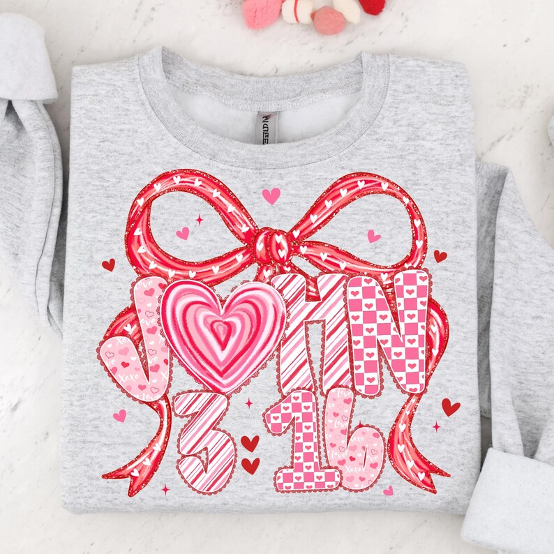 May include: A light gray sweatshirt with a large red bow and the text "JOHN 3:16" in pink and white. The design includes hearts and a heart-shaped candy. The sweatshirt is a Valentine's Day themed item.