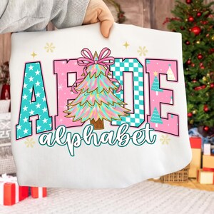May include: White sweatshirt with the word "ABCDE" in a colorful design. The letters are in teal with white stars, pink, and teal checkered patterns. A Christmas tree graphic is in the center, with the word "alphabet" below.