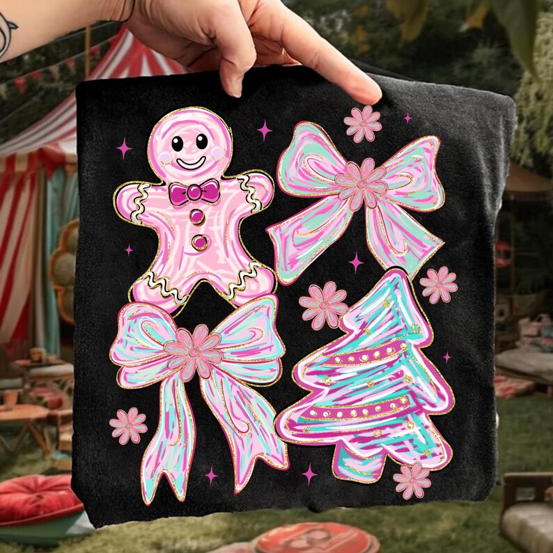 May include: Black t-shirt with a festive design featuring a gingerbread man, bows, and a Christmas tree in pink, blue, and gold. The design includes stars and flowers.