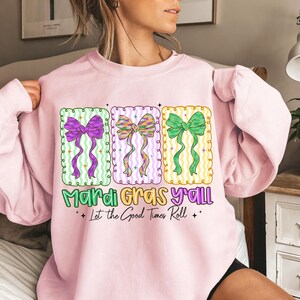 May include: A light pink sweatshirt with three rectangular designs featuring colorful bows. The text "Mardi Gras Y'all" and "Let the Good Times Roll" are printed below the designs. The bows are purple, multi-colored, and green.
