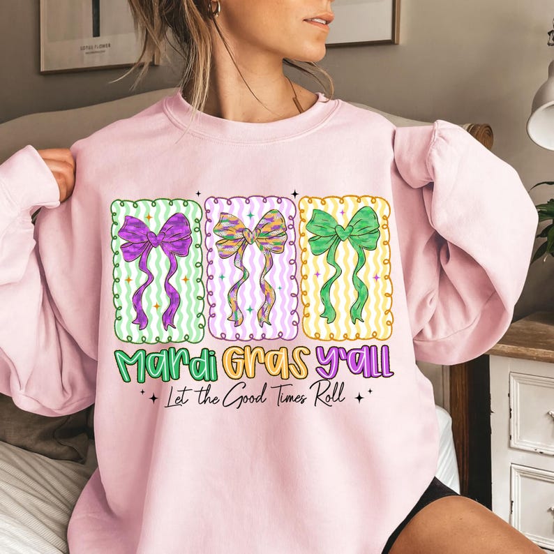 May include: A light pink sweatshirt with three rectangular designs featuring colorful bows. The text "Mardi Gras Y'all" and "Let the Good Times Roll" are printed below the designs. The bows are purple, multi-colored, and green.