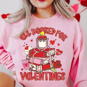 May include: Pink sweatshirt with a Valentine's Day graphic. The graphic features a stack of books, a candle, a rose, and the text "All Booked for Valentines". Hearts and a coffee cup are also included in the design.