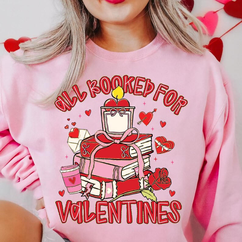 May include: Pink sweatshirt with a Valentine's Day graphic. The graphic features a stack of books, a candle, a rose, and the text "All Booked for Valentines". Hearts and a coffee cup are also included in the design.