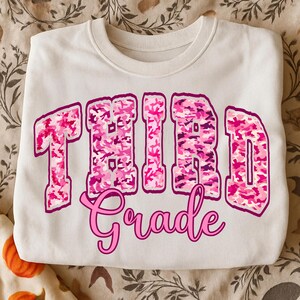 May include: A white crewneck sweatshirt with the words "THIRD Grade" in pink camouflage lettering. The word "Grade" is in a cursive font. The sweatshirt is folded and resting on a patterned surface.