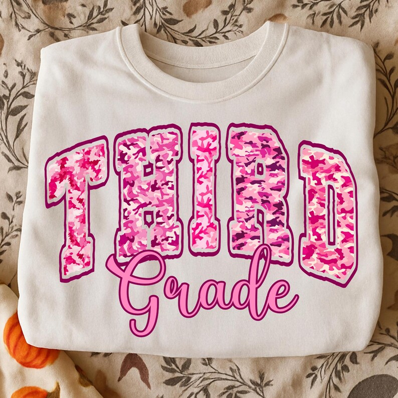 May include: A white crewneck sweatshirt with the words "THIRD Grade" in pink camouflage lettering. The word "Grade" is in a cursive font. The sweatshirt is folded and resting on a patterned surface.