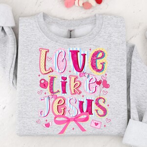 May include: A light gray sweatshirt with the words "LOVE like JESUS" in colorful, whimsical lettering. The design includes hearts, a lock, and a pink bow. The sweatshirt is folded.
