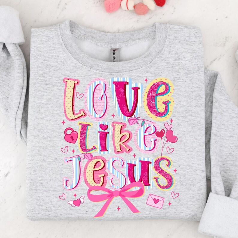 May include: A light gray sweatshirt with the words "LOVE like JESUS" in colorful, whimsical lettering. The design includes hearts, a lock, and a pink bow. The sweatshirt is folded.
