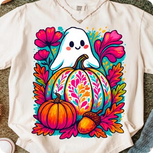 May include: White t-shirt featuring a colorful graphic of a friendly ghost, pumpkins, and autumn leaves. The design includes a large pumpkin with floral patterns, a smaller pumpkin, and an acorn. The shirt has a Comfort Colors tag.