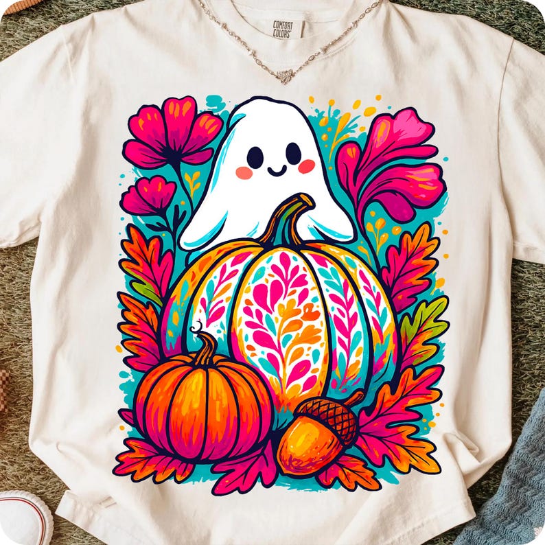 May include: White t-shirt featuring a colorful graphic of a friendly ghost, pumpkins, and autumn leaves. The design includes a large pumpkin with floral patterns, a smaller pumpkin, and an acorn. The shirt has a Comfort Colors tag.