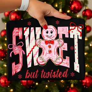 May include: Black sweatshirt featuring the words "SWEET" and "but twisted" in red and pink. A pink gingerbread man with a red bow tie is in the center. Pink bows and snowflakes decorate the design.