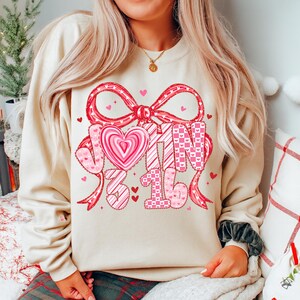 May include: A cream-colored sweatshirt with a large Valentine's Day design. The design includes a red bow, hearts, and the text "JOIN 3 16" in pink and red. The sweatshirt is a casual, comfortable style.