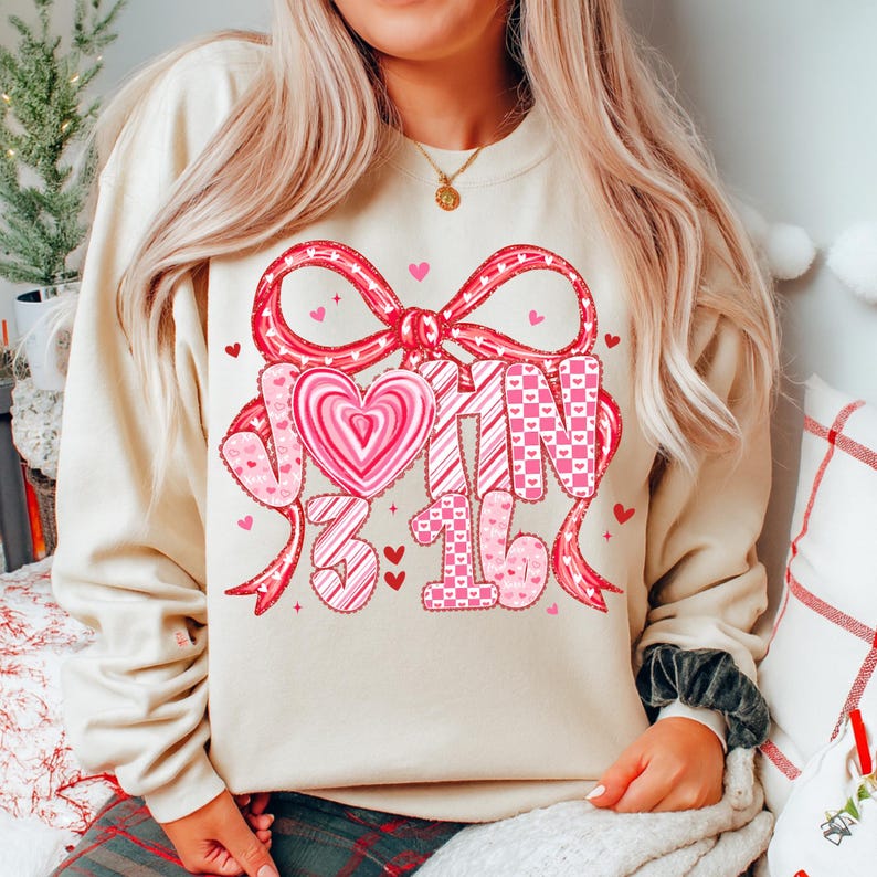 May include: A cream-colored sweatshirt with a large Valentine's Day design. The design includes a red bow, hearts, and the text "JOIN 3 16" in pink and red. The sweatshirt is a casual, comfortable style.