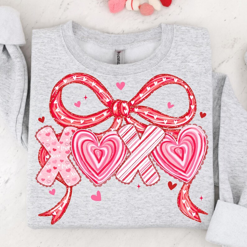 May include: A light grey sweatshirt with a large red bow and the letters "XOX" with heart designs. The hearts and bow are red and pink, with small pink hearts scattered around the design. The sweatshirt has a Valentine's Day theme.