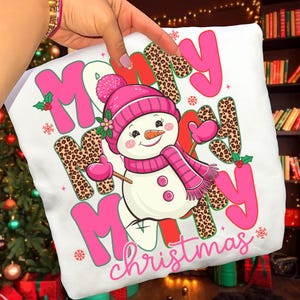 May include: White pillow with a snowman graphic wearing a pink hat, scarf, and mittens. The words "Merry Christmas" are in pink, red, and leopard print. Festive holiday decor.