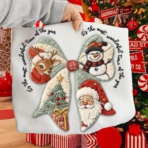 May include: A white Christmas-themed pillow with a bow design. The bow features embroidered images of a reindeer, snowman, Christmas tree, Santa Claus, and a gift. The text "It's the most wonderful time of the year" is written around the bow.
