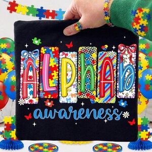 May include: Black t-shirt with the word "ALPHA" in colourful puzzle piece letters and the word "awareness" in blue script. The shirt is held up by a hand wearing a puzzle piece ring and bracelet. Balloons and decorations with puzzle piece designs are in the background.