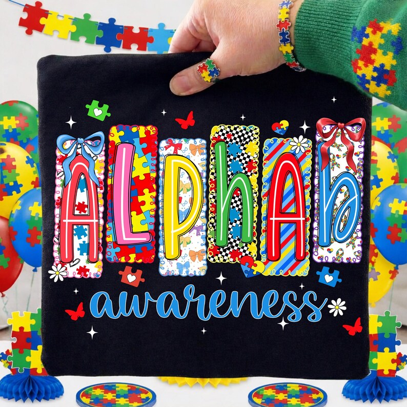 May include: Black t-shirt with the word "ALPHA" in colourful puzzle piece letters and the word "awareness" in blue script. The shirt is held up by a hand wearing a puzzle piece ring and bracelet. Balloons and decorations with puzzle piece designs are in the background.