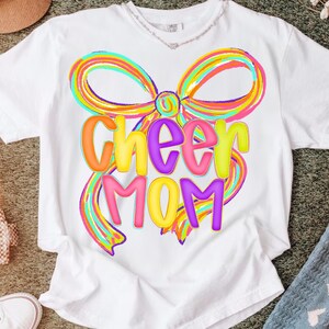 May include: White t-shirt featuring a colorful bow design with the words "Cheer Mom" in vibrant, bubble letters. The design incorporates shades of orange, yellow, green, blue, and purple, creating a cheerful and eye-catching look. A casual and comfortable apparel item.