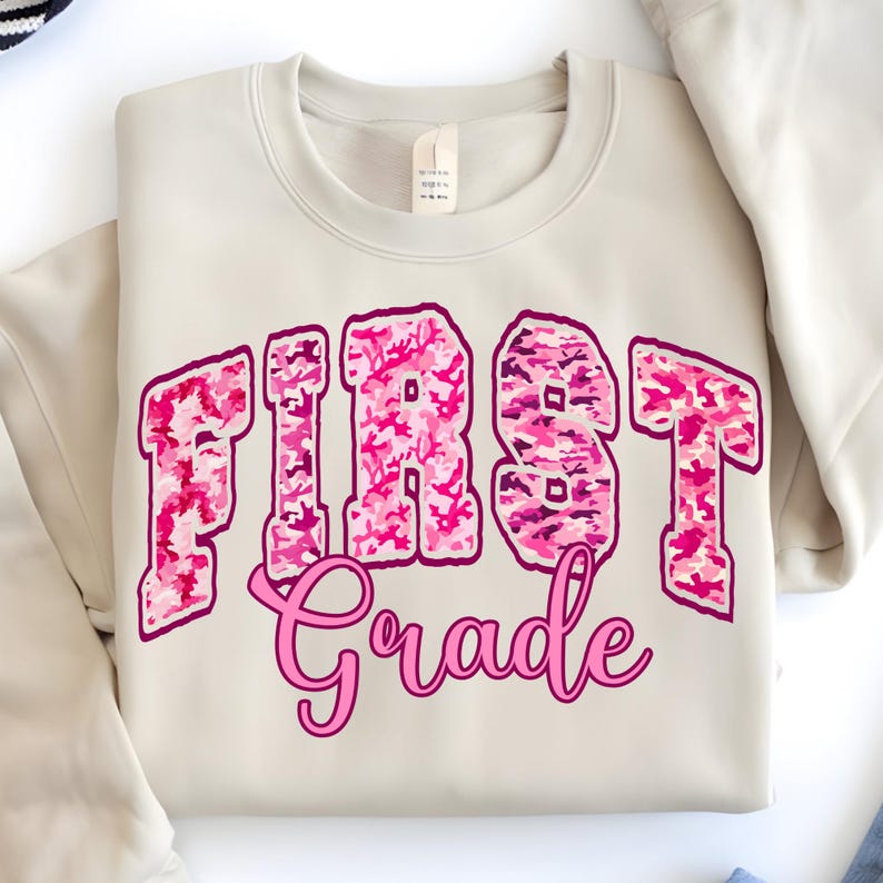 May include: A beige sweatshirt with the words "FIRST Grade" in a pink camouflage design. The word "Grade" is in a cursive font. The sweatshirt is folded neatly on a white surface.