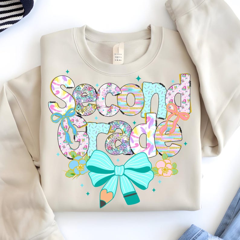May include: A beige sweatshirt with the words "Second Grade" in a colorful, whimsical design. The text features floral patterns, bows, and a pencil, with accents of turquoise, pink, and yellow. The design is centered on the front of the sweatshirt.