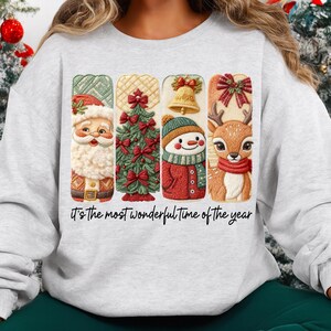 May include: A light gray sweatshirt featuring a festive design with Santa Claus, a Christmas tree, a snowman, and a reindeer. The design includes the text "It's the most wonderful time of the year".