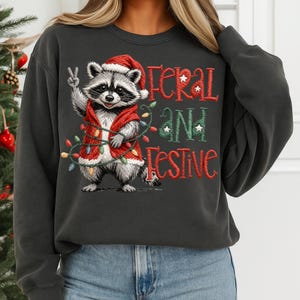May include: Dark gray sweatshirt featuring a raccoon wearing a Santa hat and vest, holding up a peace sign. The text "Feral and Festive" is embroidered in red and green. A string of Christmas lights is wrapped around the raccoon.