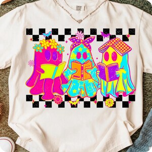 May include: Cream-colored t-shirt featuring a graphic of three cartoon ghosts reading books. The ghosts are in bright colors with floral and star accents, set against a black and white checkered background.