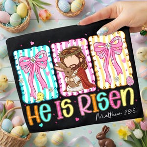 May include: Black t-shirt with colourful Easter graphics. Features illustrations of Jesus holding a bunny, pink bows, and the text "He Is Risen" with "Matthew 28:6". Decorative eggs and chicks surround the shirt.