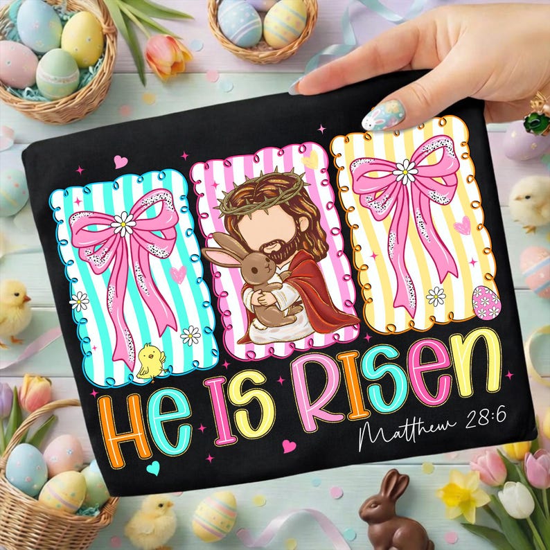 May include: Black t-shirt with colourful Easter graphics. Features illustrations of Jesus holding a bunny, pink bows, and the text "He Is Risen" with "Matthew 28:6". Decorative eggs and chicks surround the shirt.