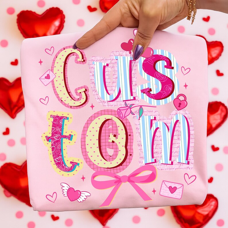 May include: A light pink fabric item with the word "CUSTOM" in a colorful, decorative font. The letters have various patterns, including stripes, hearts, and floral designs. Pink bow and heart accents complete the design.