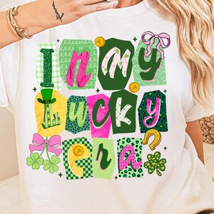 May include: White t-shirt with a colorful graphic design that says "I'm my lucky era." The design includes green, pink, and yellow elements, such as shamrocks, bows, and a horseshoe. The shirt is perfect for St. Patrick's Day.