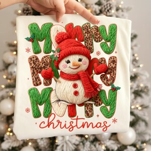 May include: Off-white Christmas-themed pillow with the words "Merry Christmas" in green, leopard print, and red. A snowman wearing a red hat, scarf, and mittens is in the center. Snowflakes and holly accents complete the design.