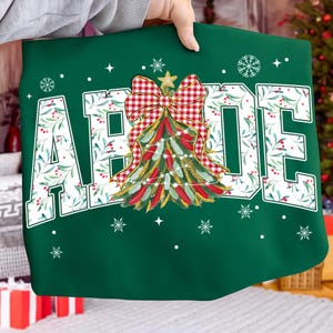 May include: A green Christmas-themed sweatshirt with the word "ABODE" in large white letters. The letters are filled with a floral pattern. A Christmas tree design with a red and white checkered bow is in the center, with snowflakes and stars.