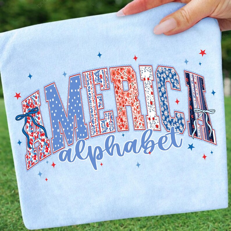May include: Light blue t-shirt with the word "AMERICA" in a decorative font, featuring red, white, and blue patterns. The word "alphabet" is written below in a script font. Small stars and bows accent the design, creating a patriotic theme.