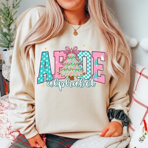 May include: Beige sweatshirt with a colorful graphic. The graphic spells out "ABCDE" with a Christmas tree in the center. The letters are in teal with white stars and pink with a checkered pattern. The word "alphabet" is written below.