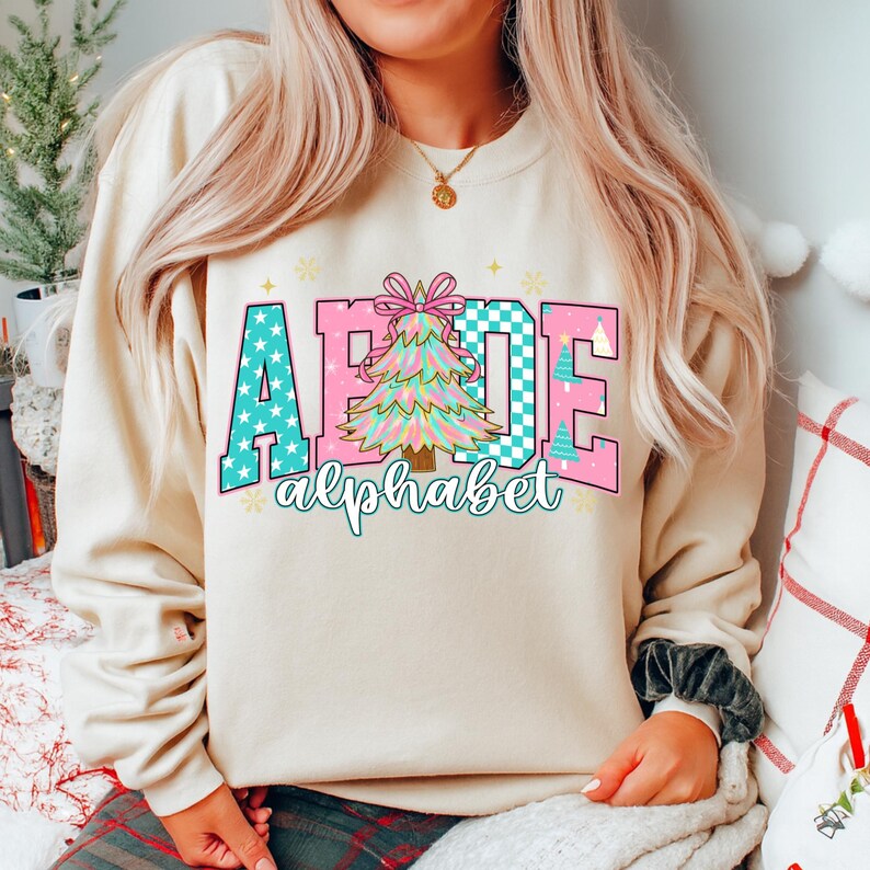 May include: Beige sweatshirt with a colorful graphic. The graphic spells out "ABCDE" with a Christmas tree in the center. The letters are in teal with white stars and pink with a checkered pattern. The word "alphabet" is written below.