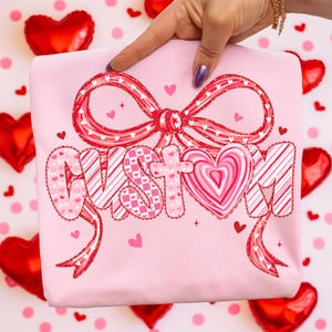 May include: A light pink sweatshirt featuring a large bow and the word "CUSTOM" in a decorative font. The bow and letters are in shades of red and pink with heart patterns. Small hearts are scattered around the design.