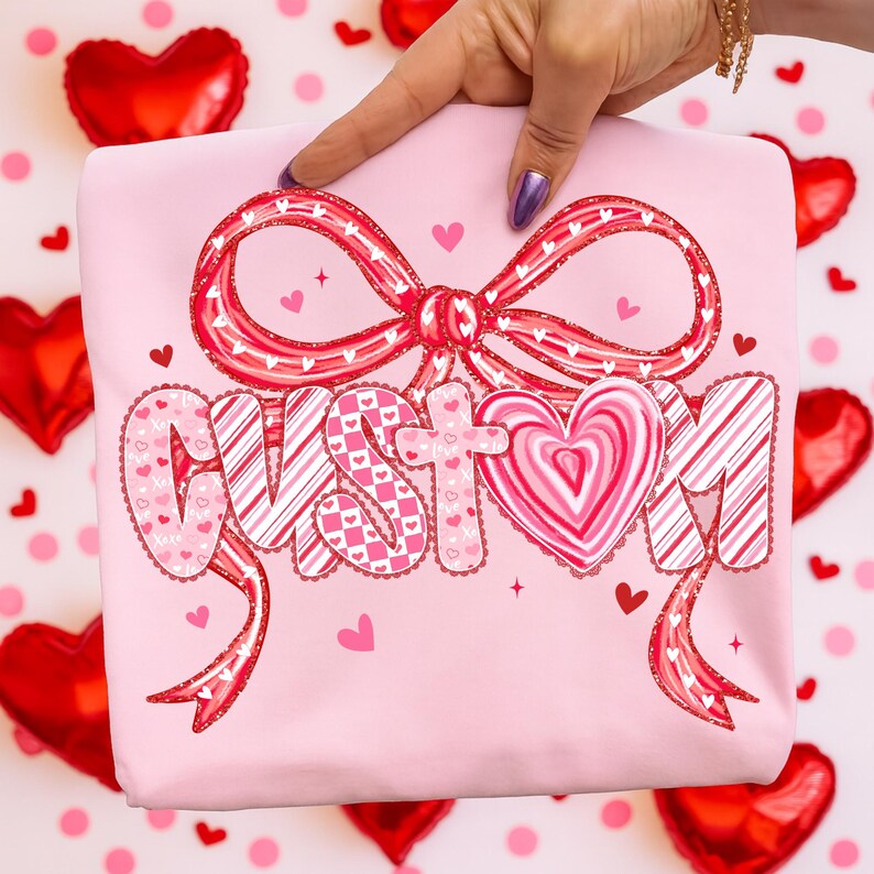 May include: A light pink sweatshirt featuring a large bow and the word "CUSTOM" in a decorative font. The bow and letters are in shades of red and pink with heart patterns. Small hearts are scattered around the design.
