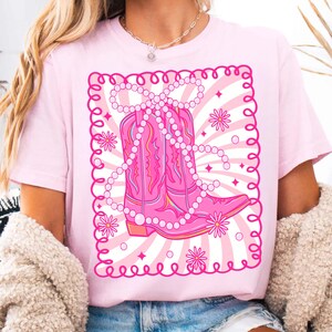 May include: A light pink t-shirt featuring a graphic of a pink cowboy boot adorned with a pearl necklace and floral accents. The design is set against a backdrop of white and pink rays and a decorative border. The shirt is a solid light pink color.