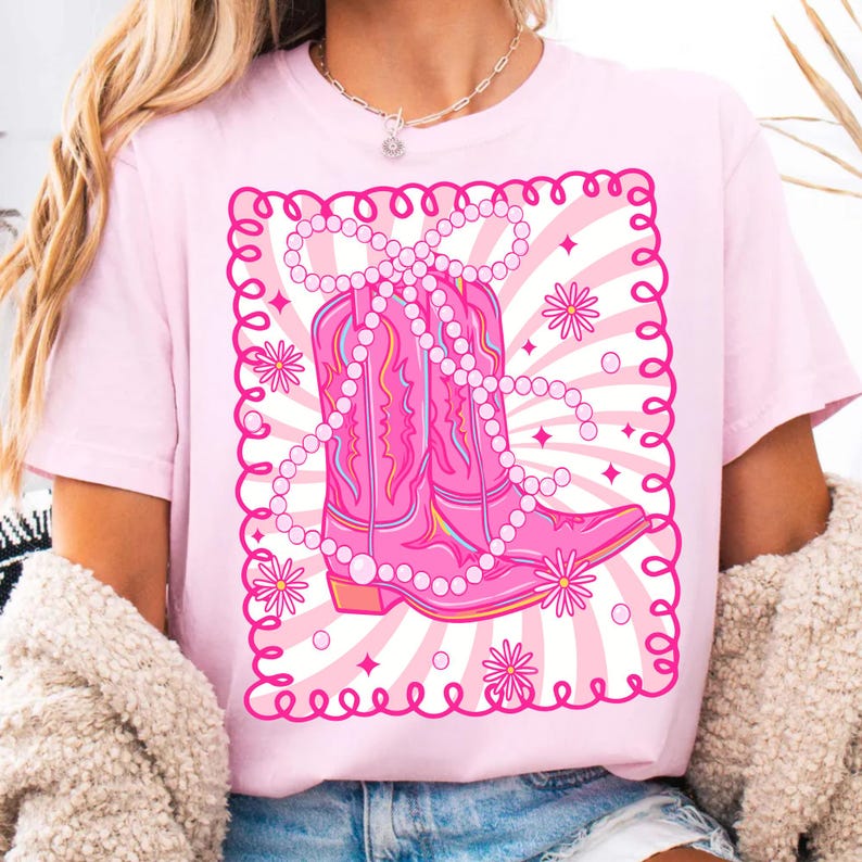 May include: A light pink t-shirt featuring a graphic of a pink cowboy boot adorned with a pearl necklace and floral accents. The design is set against a backdrop of white and pink rays and a decorative border. The shirt is a solid light pink color.