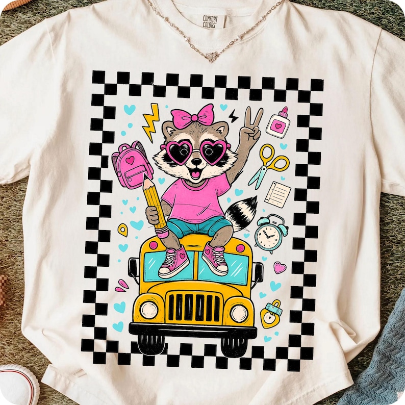May include: White t-shirt with a colorful graphic of a raccoon wearing heart-shaped sunglasses and a pink bow, sitting on a yellow school bus. The raccoon holds a pencil and makes a peace sign. The design includes school-themed elements and a black and white checkered border.