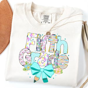Op de afbeelding: Cr&egrave;mekleurig T-shirt met de tekst "Fifth Grade" in een kleurrijk patroon. Het ontwerp bevat een lichtblauwe strik met een potlood, bloemen en kleine blauwe sterren. Een gouden ketting is over het shirt gedrapeerd.