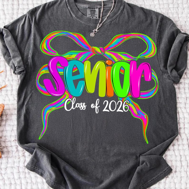 May include: Dark grey t-shirt featuring a vibrant bow design and the word "Senior" in multi-coloured letters. The text "Class of 2026" is below. A casual, comfortable style of shirt.